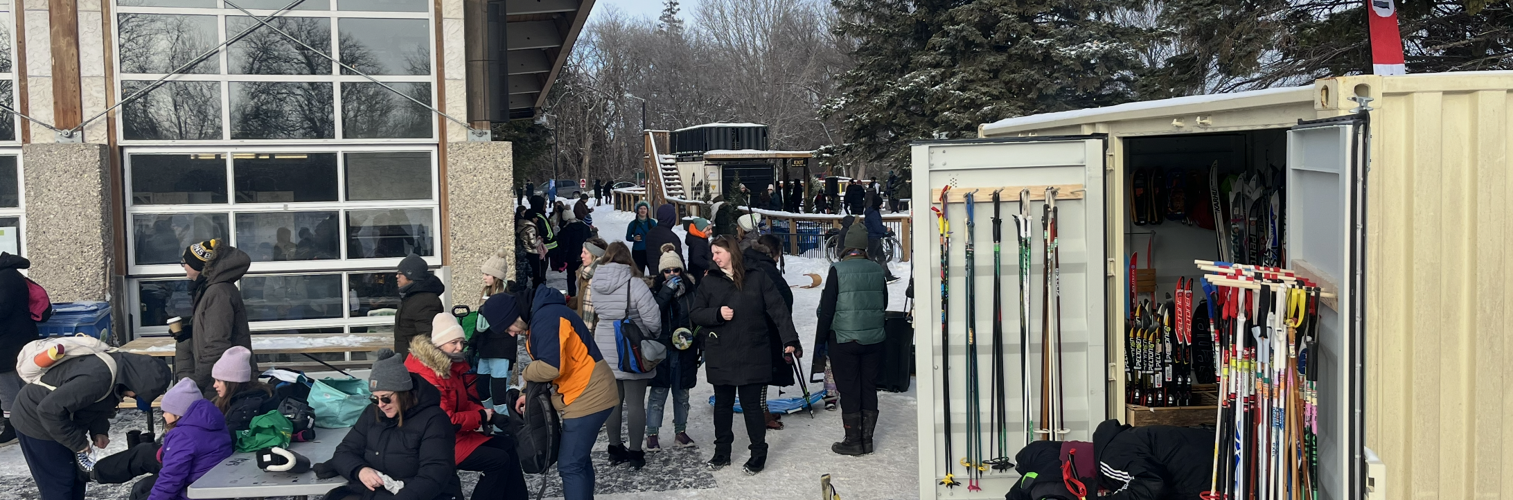 A shipping container full of ski equipment beside a glass building with a large crowd of people in winter